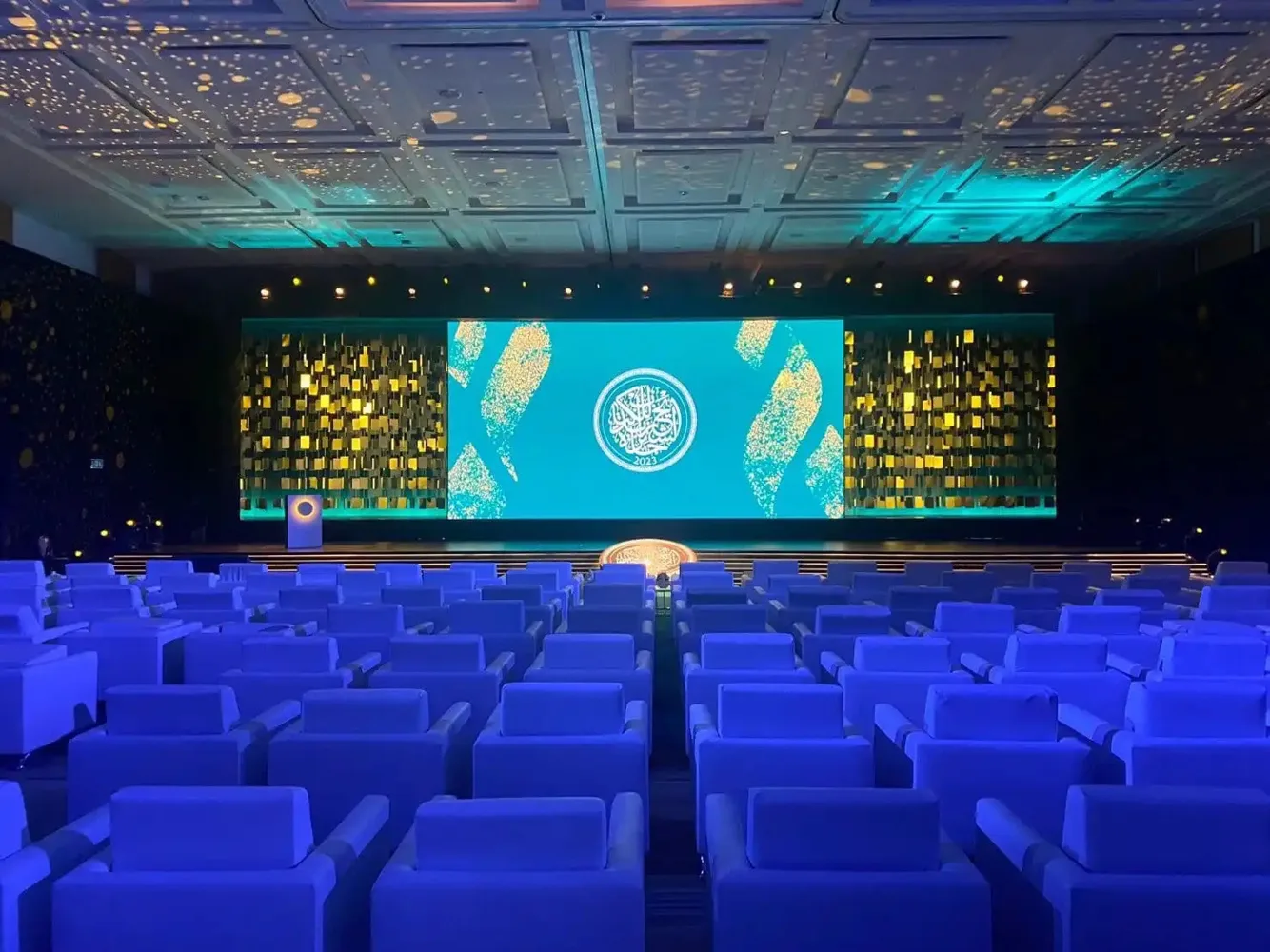Conference venue with a lit stage, large LED screen and blue auditorium lighting